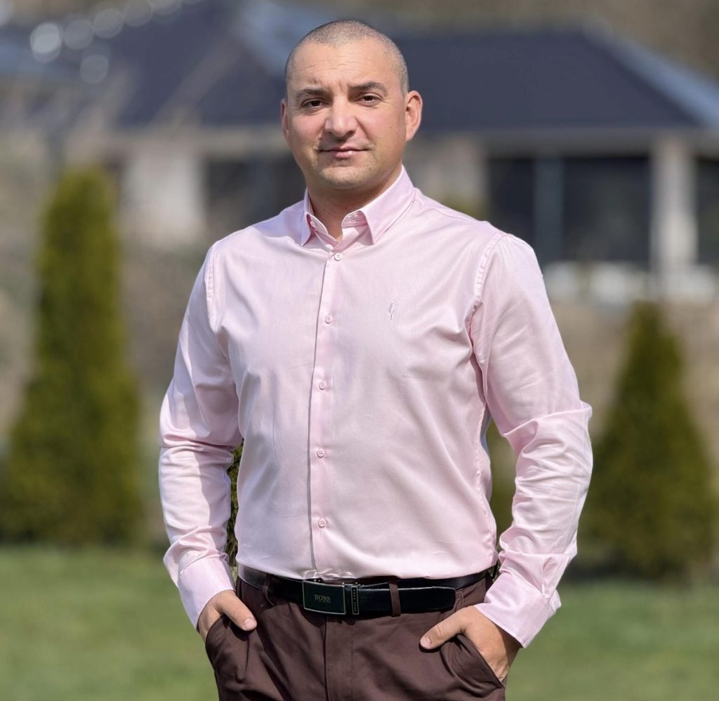 Mężczyzna w różowej koszuli i brązowych spodniach na trawniku przed domem.